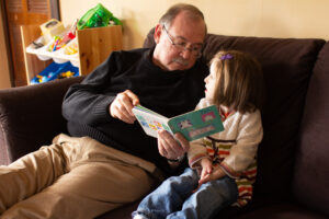 Jack (Sarah and Amy's grandfather) in a picture taken 15 years ago. He is sitting on a sofa reading Moo, Baa, La, La, La with Sarah. They are looking at each other and it seems like they are at a dramatic moment in the book.