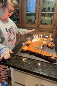 Sarah in a gray sweatshirt with a tiger printed on the front. She is standing at a black kitchen counter next to a tiger cake with lit candles. She looks excited.