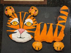 Orange, black, and white tiger cake. The head is one large circle cake. The ears, body, tail, and paws are cupcakes covered with fondant.