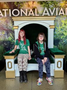 Amy and Sarah in matching green shirts that say “I’ve got your back Amy” are sitting on a bench with a sign above them for the National Aviary