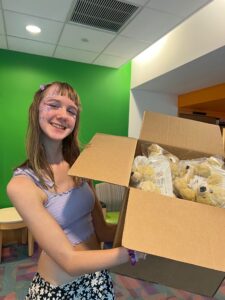 Amy has her long blond hair down and is standing in front of a green wall while wearing a lavender crop top and holding a large cardboard box tipped towards the camera so you can see it is full of bags of stuffed animal bears wearing braces. Amy is smiling.