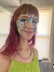 Amy in a green tank top and pink hair with a closeup of her face paint of blue and green swirls with glitter and gems