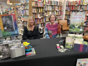 Kathleen Somers and Jennifer Celeste Briggs sit behind a table in a bookstore with piles of their books in front of them. 