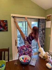 Amy in a purple and white striped sundress holding on to a curtain rod with one hand while her other hand holds a table. Her right leg is extended fully to use her toes to grab a foil wrapped egg from the open bulb end of the curtain rod