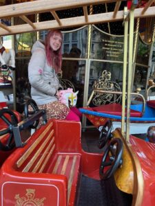 Amy a white winter coat sitting on a carousel on a very tiny horse which is in the inner circle of animals and objects. An empty firetruck is in the outer circle in front of Amy.