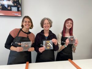 Two women (Erika and Jenny) and one teen (Amy) in a row with a white wall behind them. They are all wearing black aprons and smiling while holding chocolate watch molds that they filled and decorated