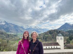 Amy in a pink sweater and Jenny in a purple fleece standing outside and smiling with a gorgeous mountain range, blue sky, and clouds behind them.