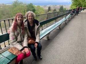 Amy and Jenny sitting in their winter coats on a very long green bench with trees and blue sky behind them