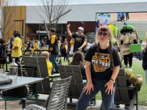 Sarah in a black t-shirt that says "412 Fest" on it. she is dancing with her hands on her legs. There are people behind her in black and gold, the Pittsburgh colors, also dancing.