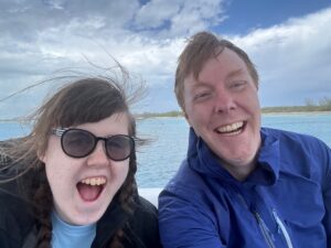 Carl and Sarah with huge smiles as their hair is whipped around by the wind. They are wearing raincoats and you can see the ocean and a coudy sky behind them