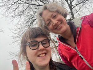 Sarah and Jenny with a closeup of their heads as if on a diagonal. They are outside with a tree behind them, smiling. Sarah is holding up two fingers.