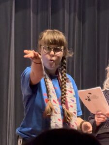 Sarah in her blue Mary Poppins shirt with her white scarf as a costume for Mrs. Banks. Her hair is in two braids. There is a black curtain behind her. She is looking directly at the audience with her arm outstretched and her mouth pursed because she has just blown a kiss.