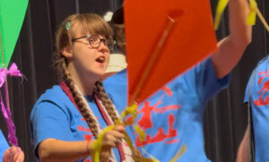 Sarah in blue shirt with red "Mary Poppins" letters. Her hair is in two braids. She is singing while holding a stick that is attached to a small red kite.