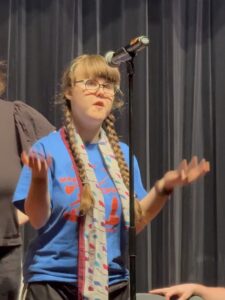 Sarah seen from the waist up with a black curtain behind her. She is on a stage and there is a microphone in a stand in front of her. She is wearing a blue t-shirt with red letters for "Mary Poppins" and she has a white scarf with blue and red squares on it. The scarf is her Mrs. Banks costume. Her elbows are bent and her hands open and palm up. Her mouth is slightly open.