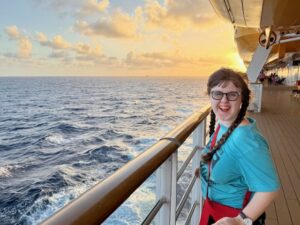 Sarah with her hair in two braids, smiling as she looks back at the camera. She is standing at the side of the ship, with the railing as high as her chin, and there are waves and a sunset in the background