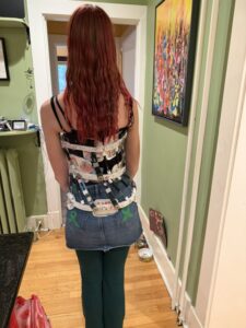 Amy is standing in the kitchen facing away from the camera. She is wearing a jean skirt and a black tank top under the Whisper brace which has white plastic pieces and metal springs and rods surrounding her torso.