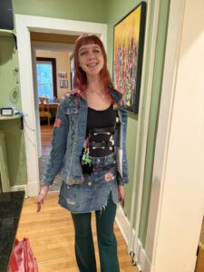 Amy is standing in a kitchen doorway, smiling with her red dyed hair framing her face. She is wearing a black shirt, a jean skirt, and a jean jacket open to show her Whisper brace attaching in the front