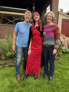Carl in a blue t-shirt and blue jeans, Amy in her sparkly red prom dress and black cloak, Jenny in a multi-colored crocheted sweater and dark blue jeans. They are standing on grass with a brick house behind them. They are smiling.