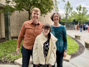 Carl in a burnt orange button down shirt and jeans, Jenny in a blue and black long sleeved blouse and dark blue jeans, Sarah in her black and gray prom dress and white fuzzy jacket. They are standing outside in front of a tree and grass, smiling. 