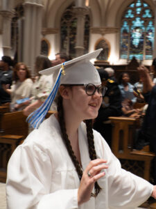 A closeup of Sarah's face as she walks down the aisle of a cathedral for graduation. She is smiling and wearing a white cap with a blue and white tassel, glasses, and a white gown.