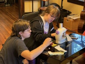 Anna and Sarah are sitting on the floor using a black piano bench as a sewing table. There is a small white sewing machine with small pieces of fabric that Sarah is guiding through the machine. Anna is helping make sure everything goes smoothly and safely. Anna is in a black t-shirt. Sarah is wearing a white t-shirt and a black unzipped hoodie.