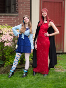 Amy's bestie in a blue dress and light blue half-sweater, white lacy tights with a fuzzy blue yarn winding around her right leg. Amy is in a full-length sparkly red dress, a black cloak, and her hair is dyed red