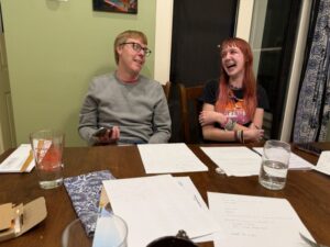 Carl in a gray sweatshirt and Amy in a black-t-shirt with a pink heart, sitting at a brown dining room table together with a green wall behind them. They are looking at each other. Carl looks like he is finishing saying something and Amy is laughing.