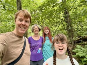 Carl in a light brown t-shirt, Jenny in a purple t-shrt, Amy in a turquoise sweatshirt, and Sarah in a white t-shirt, all smiling as they stand in the woods.