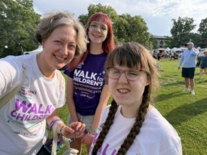 Jenny in a white shirt, Amy in a purple shirt, and Sarah in a white shirt smile in the sunshine with green trees and grass behind them. The sky is blue. They are ready for the Walk for Children's Hospital.
