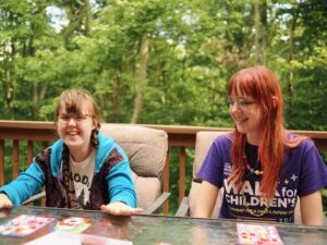 Sarah and Amy smiling as they sit at an outside table with trees in the background. Sarah is smiling and looking at the camera, wearing a turquoise sweatshirt open over a white t-shirt. She has glasses and her hair is in two braids. Amy has dyed red hair and is smiling and looking down and towards Sarah. She is wearing a purple Walk for Childrens shirt.