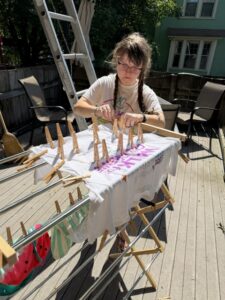 Sarah is standing in the sun next to a clothes drying rack. She is wearing a white t-shirt and her hair is in two braids. She is focusing on attaching 31 clothespins to the white shirt spread on the drying rack.