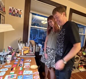 Amy and Carl standing and smiling, seen from the side, as they look down at a desk covered in small drawings of flags and potatoes that Amy made Carl for his birthday. Amy is in a flowered skirt and blue tank top. Carl is in a black t-shirt with math equations on it.