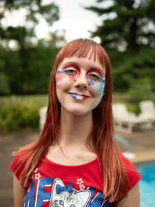 Close-up of Amy with a close-lipped smile. Her face is covered in red, white, and blue facepaint. Even her lips are red, white, and blue.