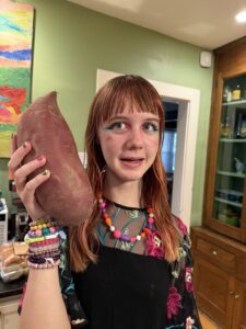 Close-up of Amy standing in the kitchen with a green wall behind her. She is holding a sweet potato up next to her head. The sweet potato is longer than her head is tall. She is looking slightly to the side at the sweet potato with a puzzled expression.