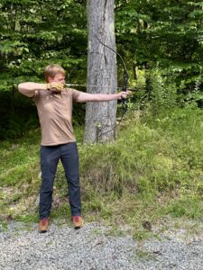 Carl in a light brown t-shirt and black pants standing with his body facing front but his face turned to the side as he pulls back the string from the bow with an arrow notched.