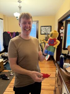Carl in a brown t-shirt, standing in foyer, smiling, holding a homemade soft stuffed LEGO version of Carl that has blue shoes, green pants, a red tank top with the number 50 on it, and removable red hair