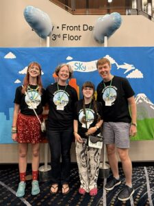 Amy, Jenny, Sarah, and Carl are wearing matching black t-shirts with pictures of the Seattle space needle. Behind them is a painted backdrop of the Seattle skyline. They are all smiling. 
