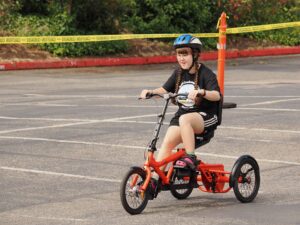 Sarah riding an orange tricycle in an empty parking lot while wearing a black t-shirt and a blue helmet and black shorts. She is smiling and looking forward. 