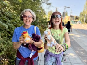 Jenny and Amy in sunglasses and t-shirts rushing up to the camera, smiling as they hold their new stuffed animals in front of them