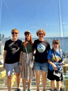 Carl, Amy, Jenny, and Sarah standing at the top of the Seattle space needle. They are outside with the glass wall behind them, so it looks like ocean and blue sky. They are smiling, all wearing t-shirts and sunglasses.