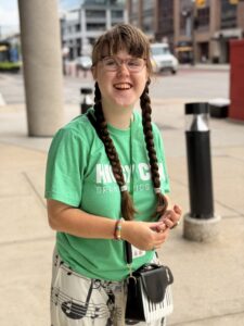 Close-up of Sarah outside with her hair in two braids. She is wearing a green Higgy Con t-shirt and smiling
