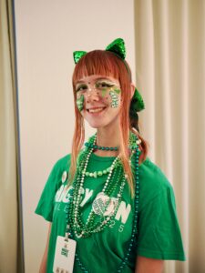 Close-up of Amy in a green Higgy Con t-shirt, sparkly green cat ears, green hair bow, green face paint designs of a spine on one cheek and a back brace on the other cheek, and many green beaded necklaces