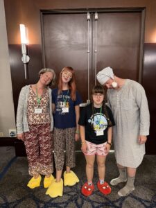 Jenny, Amy, Sarah, and Carl stand in pajamas (Carl in a long night shirt) in front of brown doors. They pretend to sleep. Carl has a blue and white striped sleeping cap with a pop-pop on the tip. Amy and Jenny have yellow goose foot slippers. Sarah has spider man slippers. 