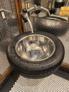 A black tire on its side with a silver sink bowl lining it and a gas pump nozzle as the sink water source