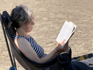 Side view of Jenny in a blue and white striped swim shirt. She is sitting in a chair on a sandy beach reading a book.