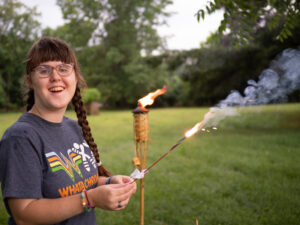 Sarah wearing a gray t-shirt, standing outside with trees behind her. She is near a tiki gas lamp and is holding a sparkler.