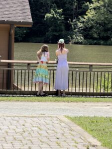 View from the back of two girls looking out over a railing at a bit of sand and greenish lake water. There is a bit of a brown wood building to the left. They are standing on grass.