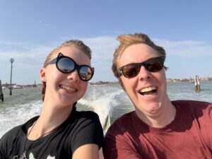 close up of Amy and Carl wearing sunglasses and smiling in the sun with the ocean behind them as they stand in a speeding water taxi. You can see the wake behind them.