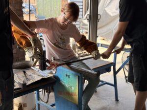 Amy is sitting on a bench wearing green leggings and a pink t-shirt and protective white leather gloves and brown leather forearm covers. She is holding a long metal rod with one hand and working with pliers to shape a glass flower that is attached to the long metal rod. You can see the arms of two helpers that are standing near her stabilizing the metal rod.
