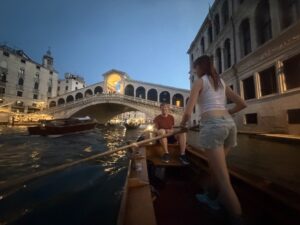 The Rialto Bridge in Venice is the main feature of the picture, at dusk. Amy and Carl are in a batela floating on the Grand Canal. Carl is sitting in the front of the boat facing the camera. Amy is facing away from the camera, standing and holding the oar that is sticking in the water and to the left of the boat. She has jean shorts and a white tank top on. There are buildings on either side of the canal.